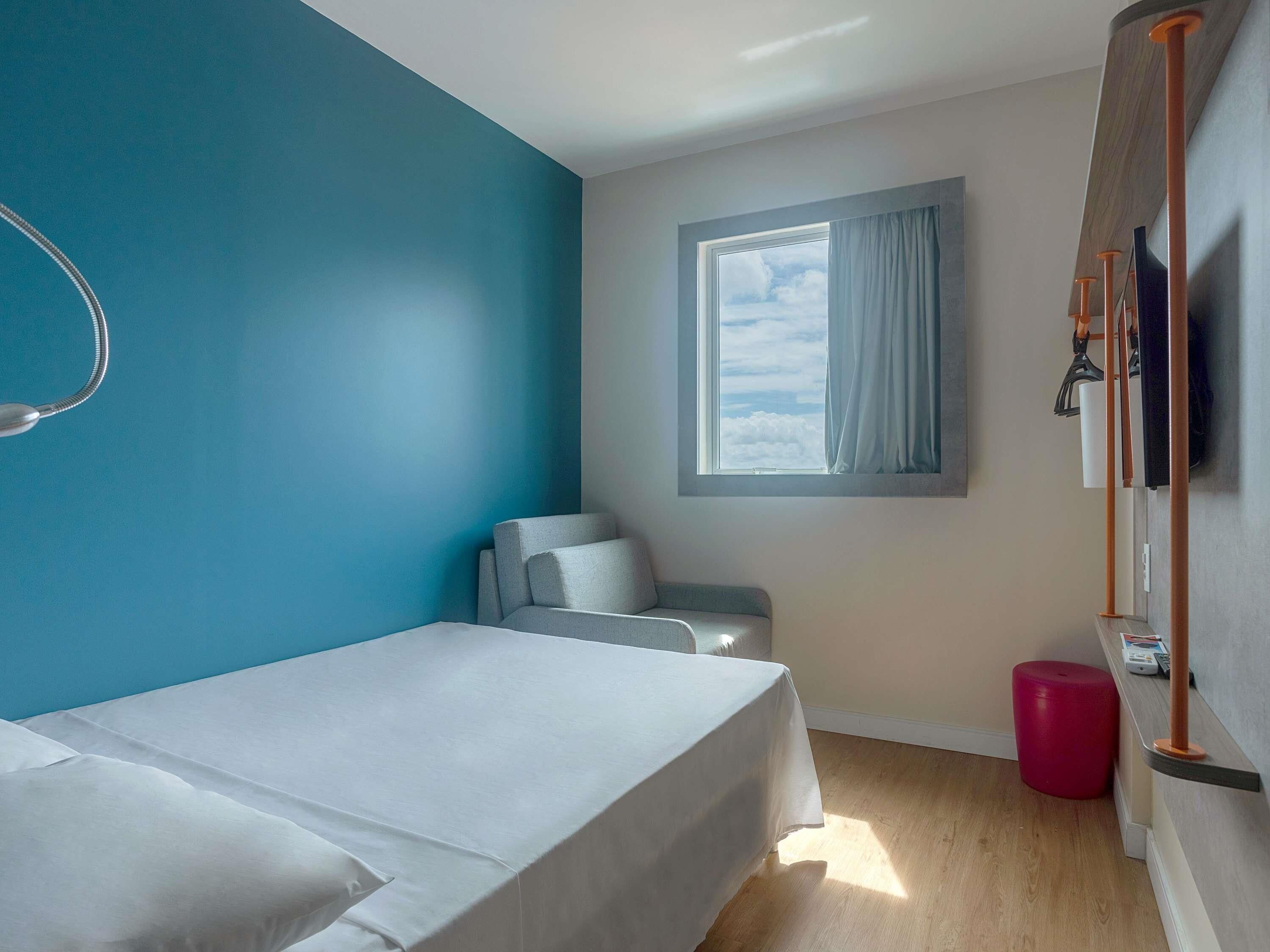 A room with a bed, a chair, a window with curtains, a red stool, and a wooden shelf with items.