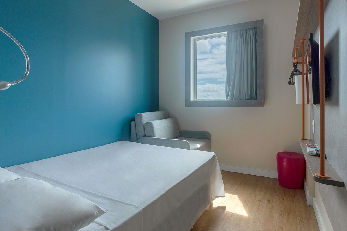 A room with a bed, a chair, a window with curtains, a red stool, and a wooden shelf with items.