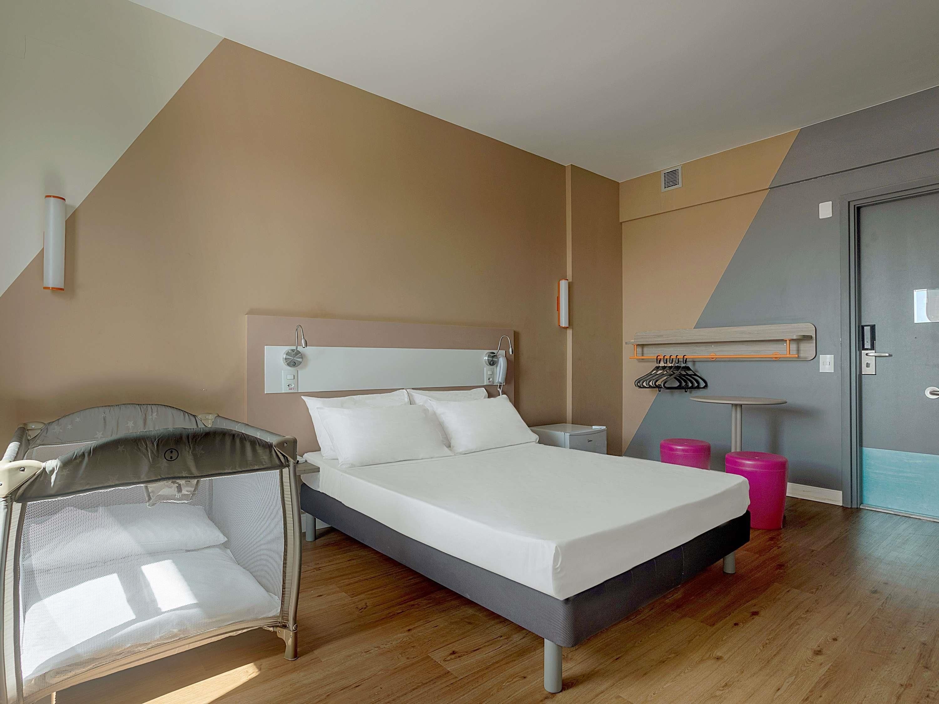 Una habitación de hotel moderna con cama, un escritorio pequeño, una silla, perchero y puerta.