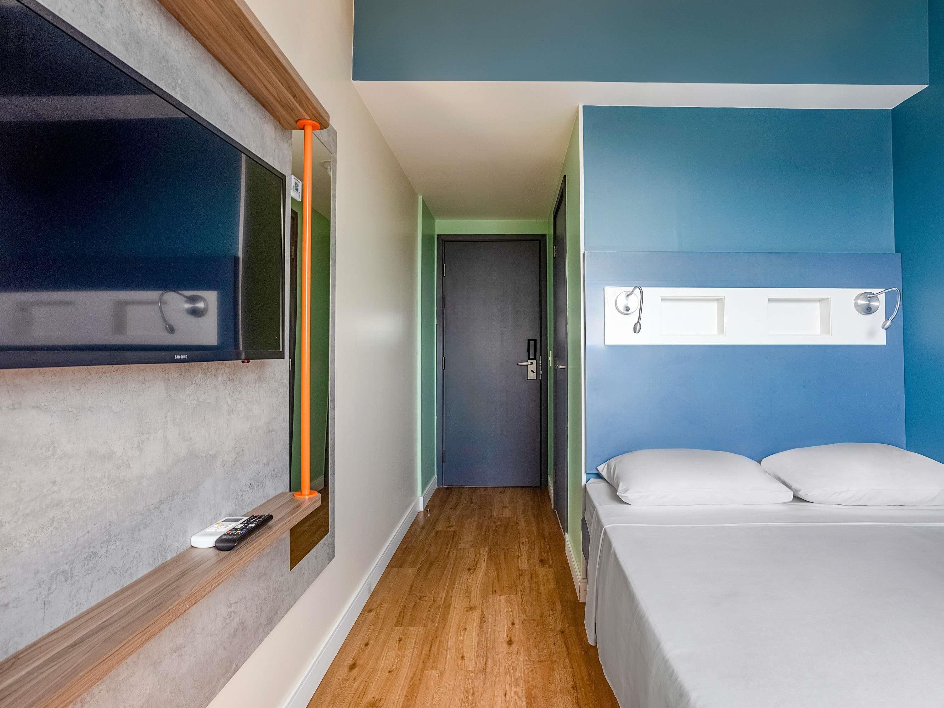 A hotel room with a bed, a TV mounted on the wall, a wooden shelf with a remote control, and a door with a handle.