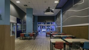 Um restaurante com várias mesas e cadeiras, um grande lustre pendurado e vista para a rua pelas janelas.