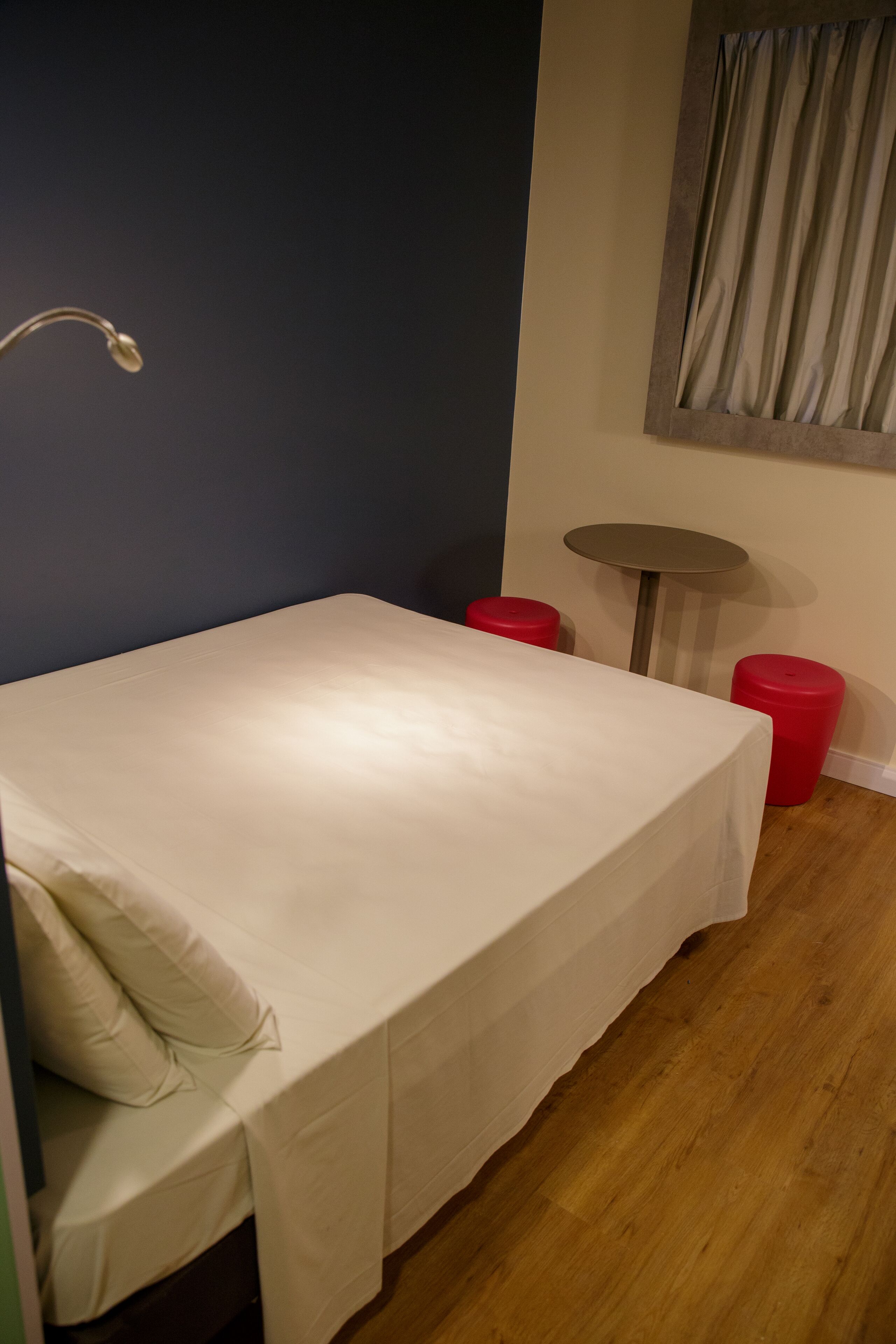 Una cama pequeña y bien arreglada con colchón y almohadas blancos, una mesita redonda y dos taburetes rojos en una habitación con suelo de madera y pared azul oscuro.