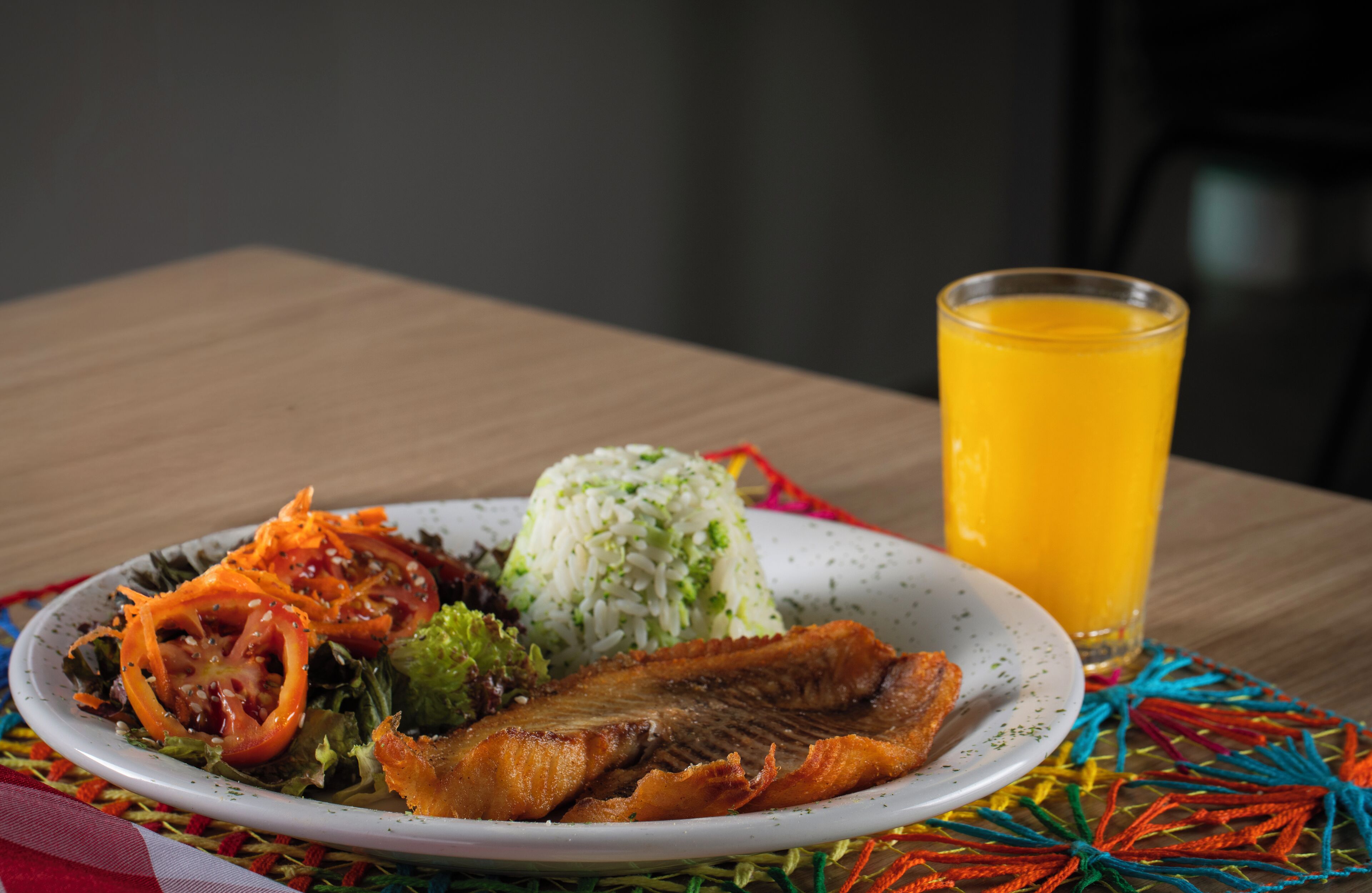 A plate of food including rice, vegetables, and a fish, with a glass of orange juice on a table.