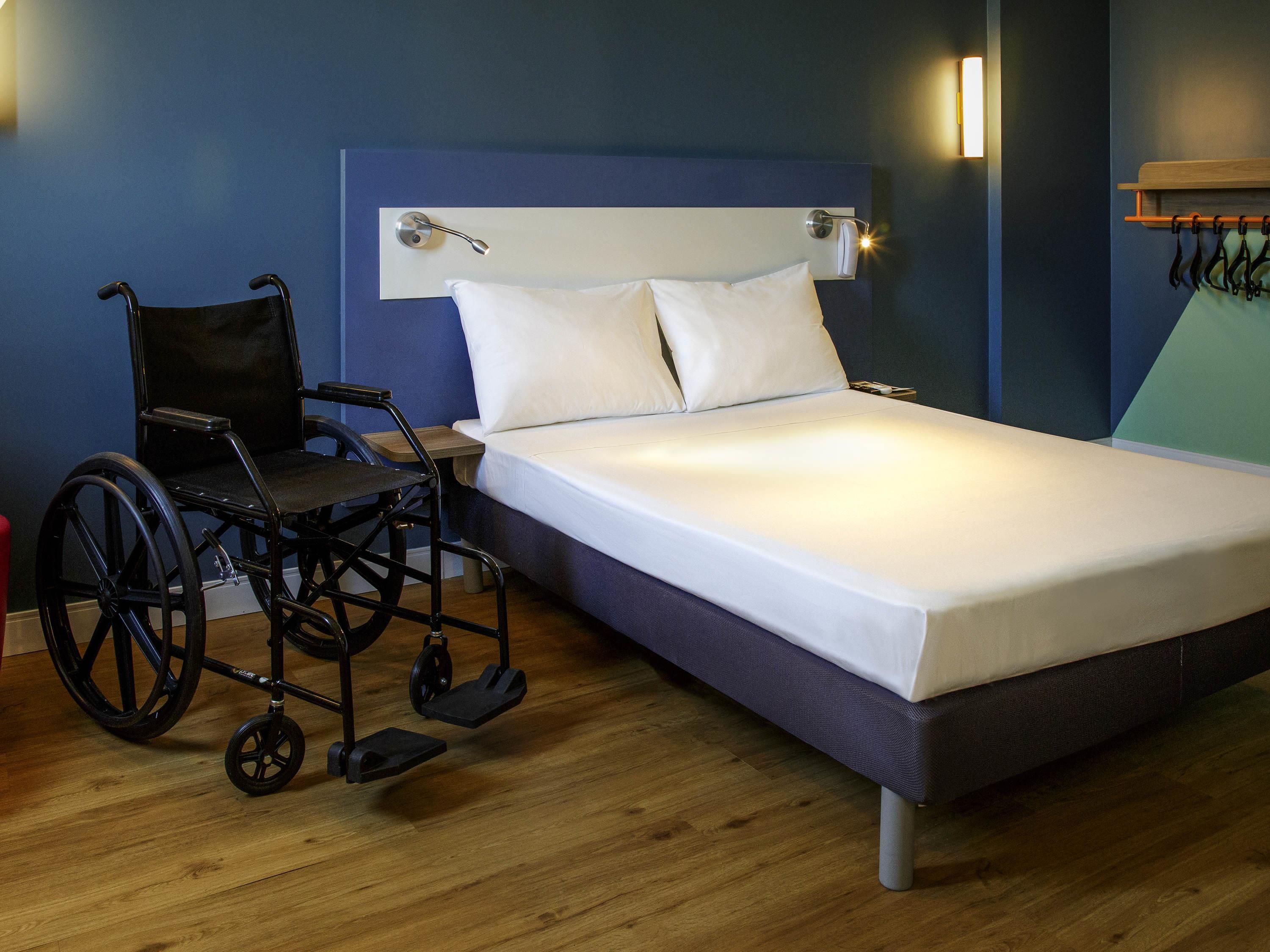 A single bed with white linens, a headboard, and a wheelchair in a room with wooden flooring and blue walls.
