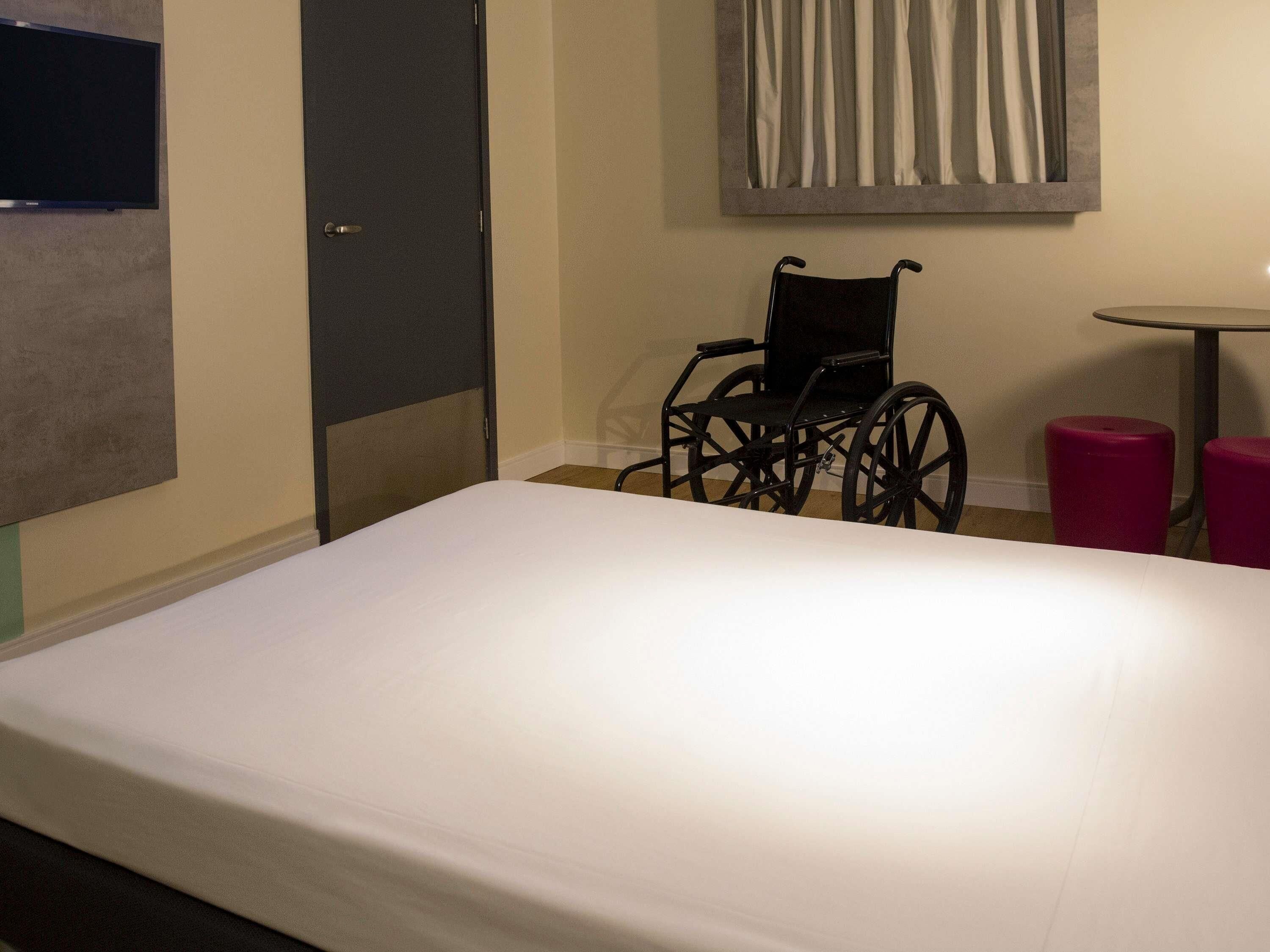 A hotel room with a bed, a wheelchair, a small table, and a chair.