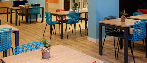Un comedor moderno con varias mesas y sillas, suelo de madera y vista a la cocina a través de una puerta de vidrio.
