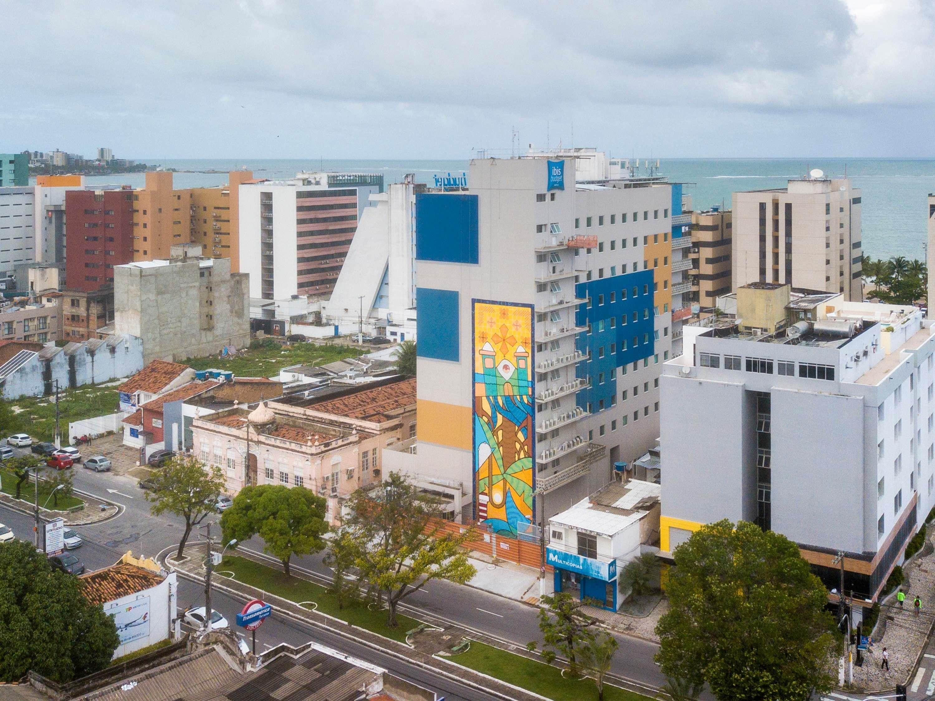 A cityscape with modern buildings, a prominent mural, and a view of the sea.