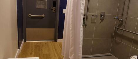A bathroom with a shower, a toilet, a towel rack, and a door with a handrail.