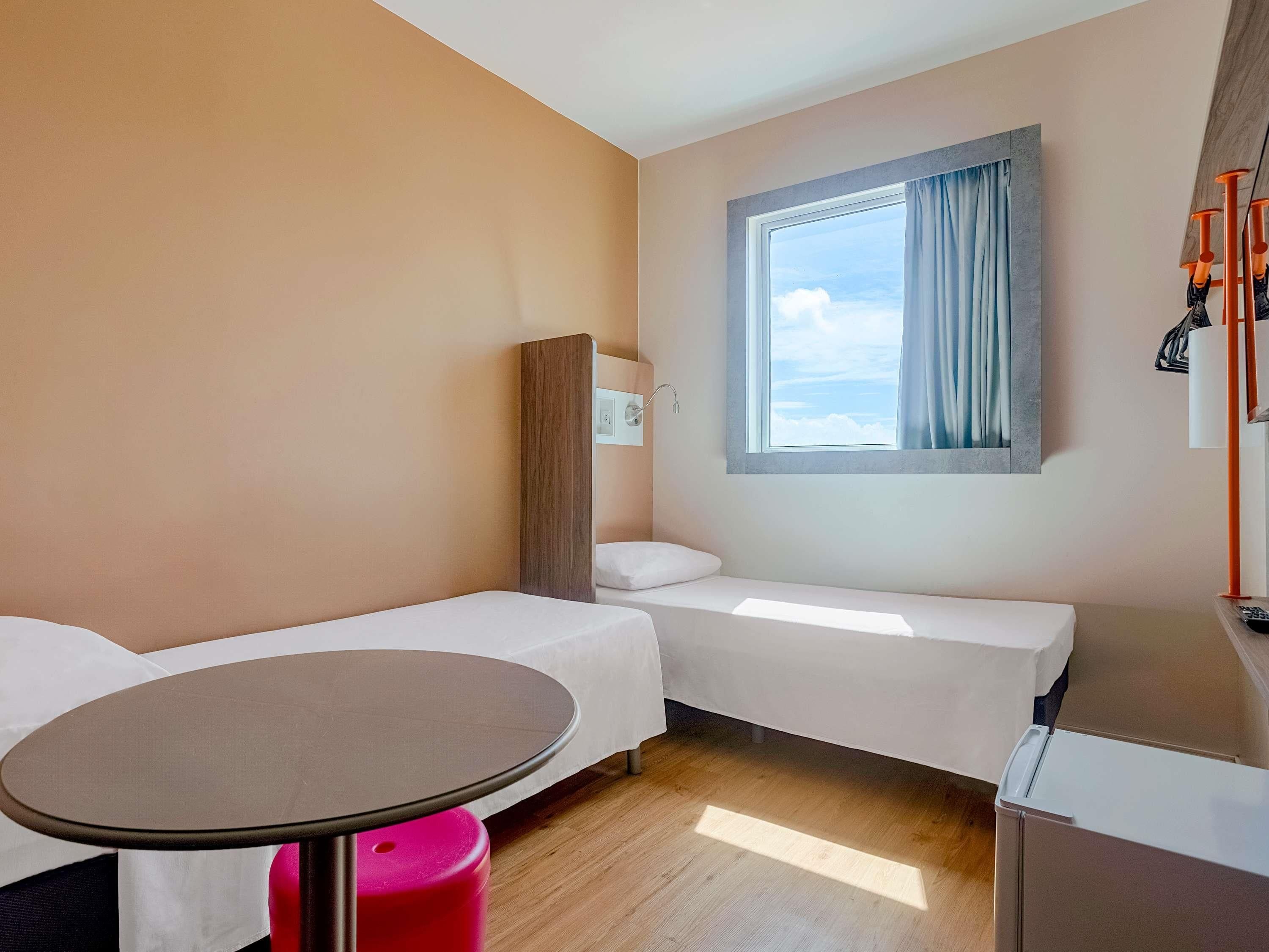 Ein Hotelzimmer mit zwei Einzelbetten, einem kleinen Tisch, einem Kleiderschrank und einem Fenster mit Blick auf den Himmel.