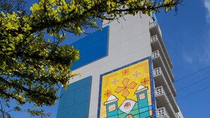 Um edifício com um mural colorido na fachada, entrada azul e branca e uma varanda visível.