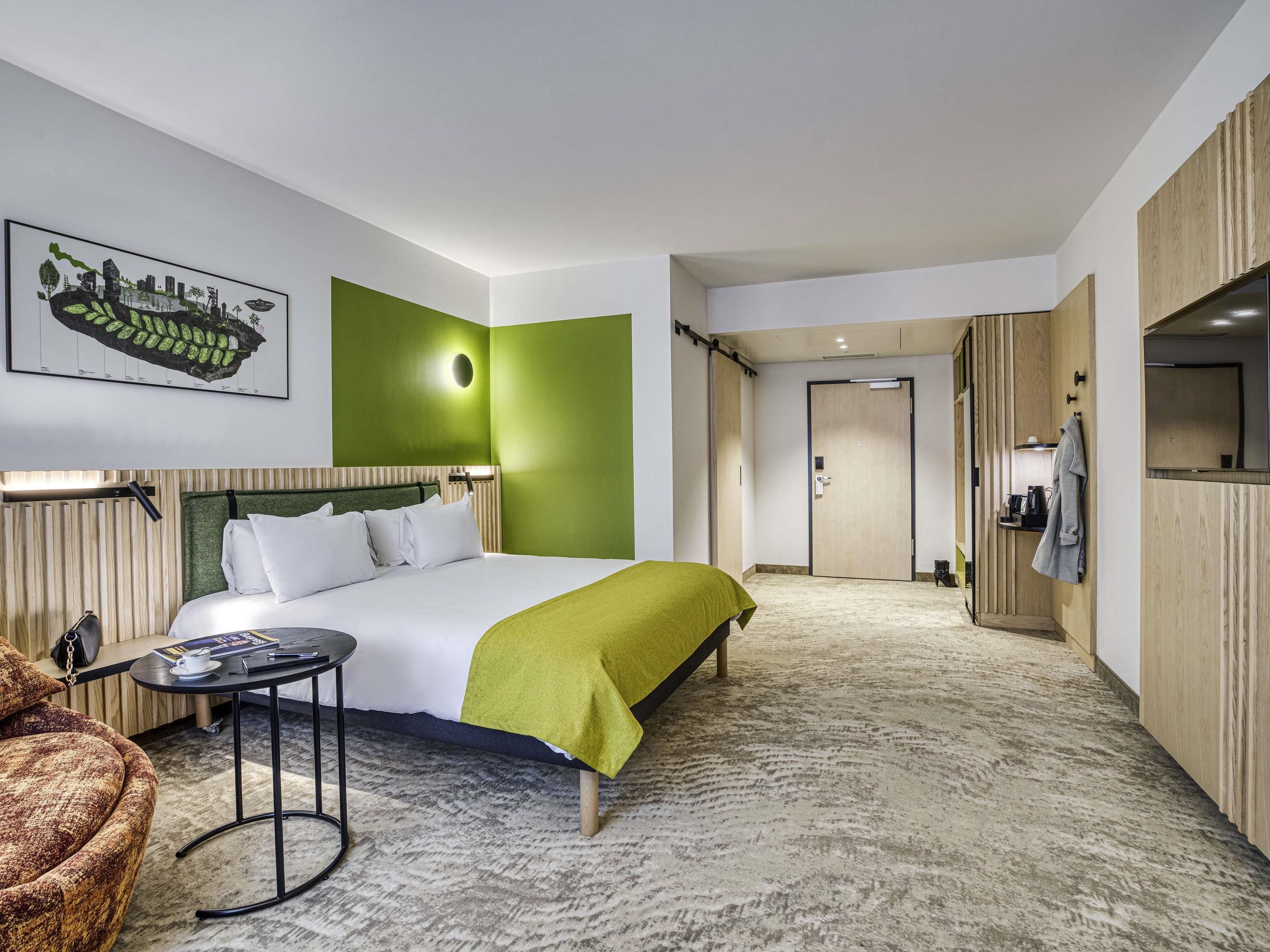Una habitación de hotel con una cama, una mesita, una silla, un perchero y una puerta que da a otra habitación.