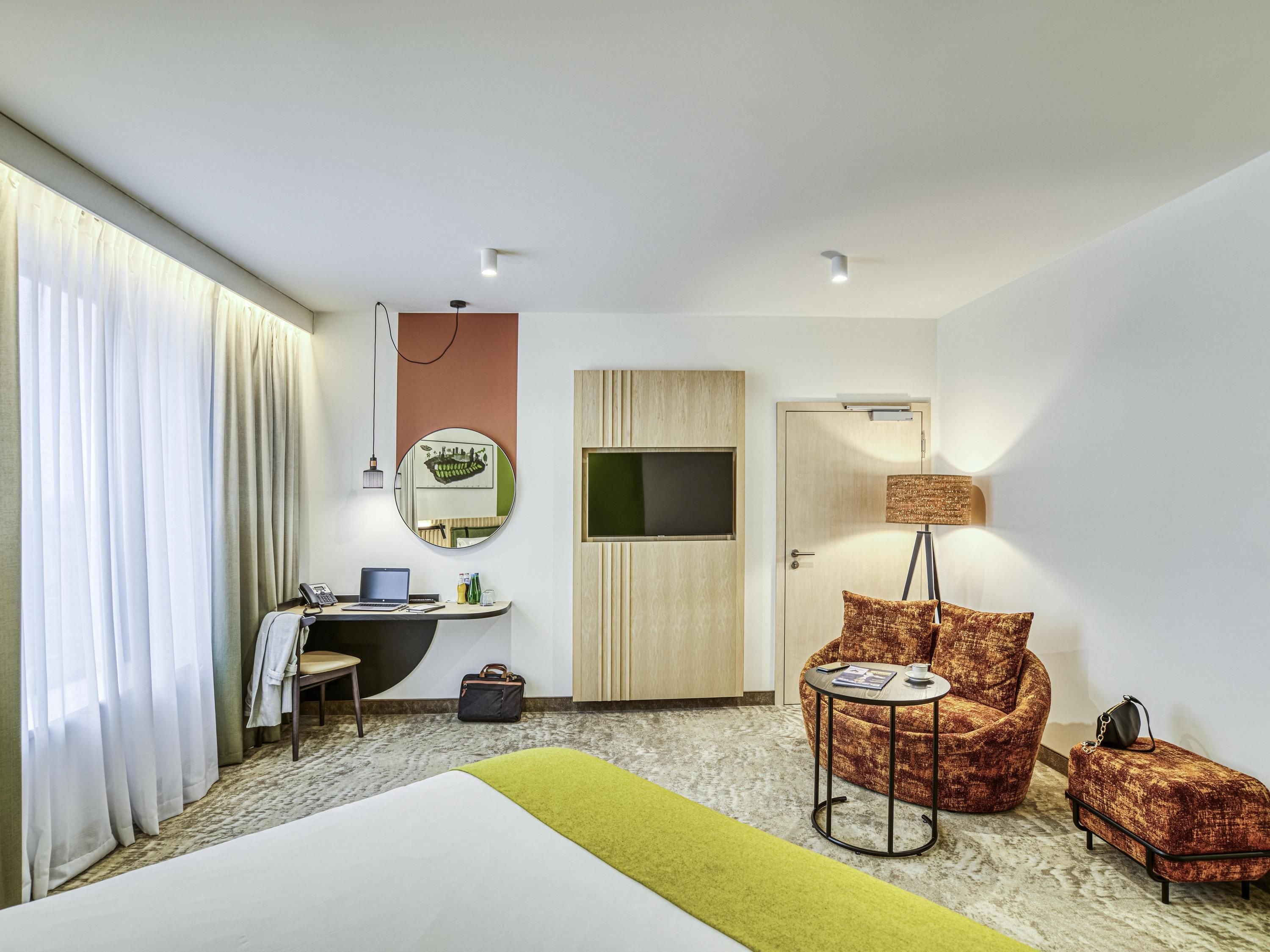 A hotel room with a bed, a desk with a laptop, a chair, a small table with a lamp, a suitcase, and a wardrobe.