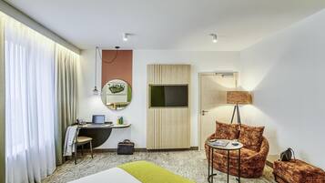 A hotel room with a bed, a desk with a laptop, a chair, a small table with a lamp, a suitcase, and a wardrobe.