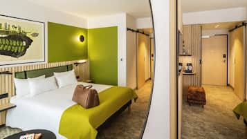 A hotel room with a bed, a bedside table, a lamp, a green wall, a framed picture, and a hallway leading to a door.