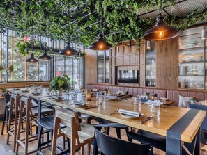 Un restaurante con una larga mesa de madera dispuesta para comer, plantas colgantes e iluminación colgante.