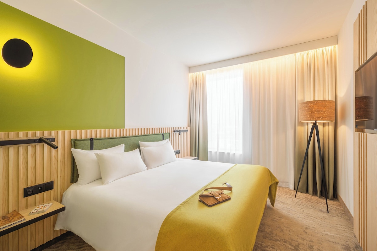 Una habitación de hotel con una cama, una mesita de noche, una lámpara y una ventana con cortinas.