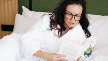 A person lying on a bed with a book and a cup, in a room with a green wall and wooden headboard.