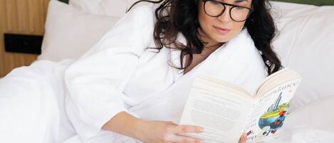 Una persona recostada en una cama con un libro y una taza, en una habitación con pared verde y cabecera de madera.