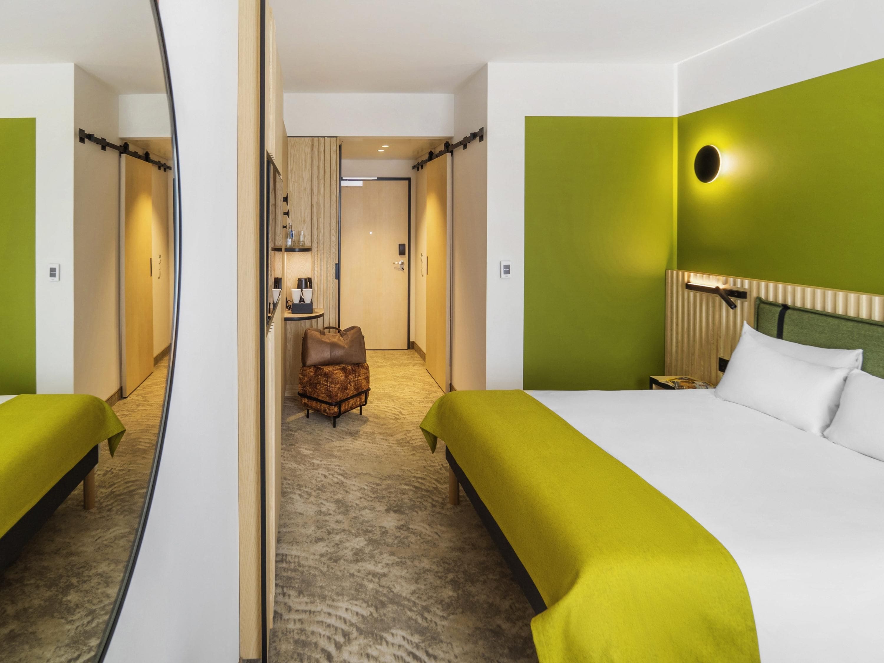 A hotel room with a large bed, a chair, and a mirror reflecting the room.