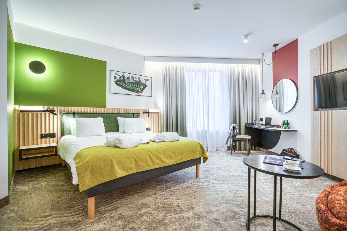 Una habitación de hotel moderna con una cama, un escritorio, una silla, un televisor y un ventanal con cortinas.