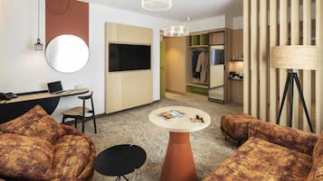 Una habitación de hotel moderna con escritorio, dos sofás, una mesa redonda de centro, televisor, espejo, armario y lámpara.