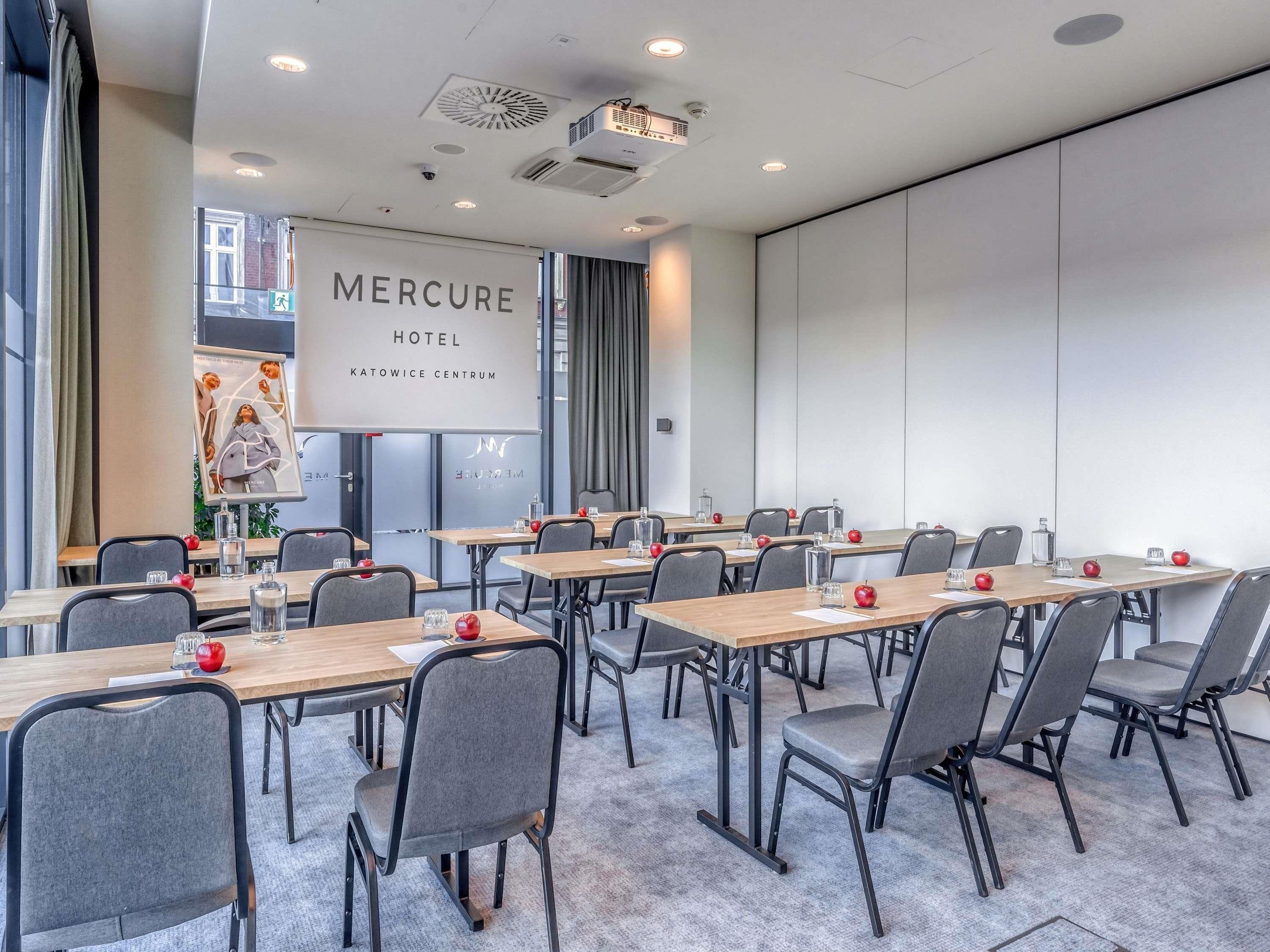A conference room with multiple tables and chairs, a large screen displaying 'Mercure Hotel Katowice Centrum', and a view of the outside through large windows.