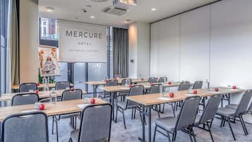 Un salón de conferencias con varias mesas y sillas, una pantalla grande mostrando 'Mercure Hotel Katowice Centrum' y vistas al exterior a través de amplios ventanales.