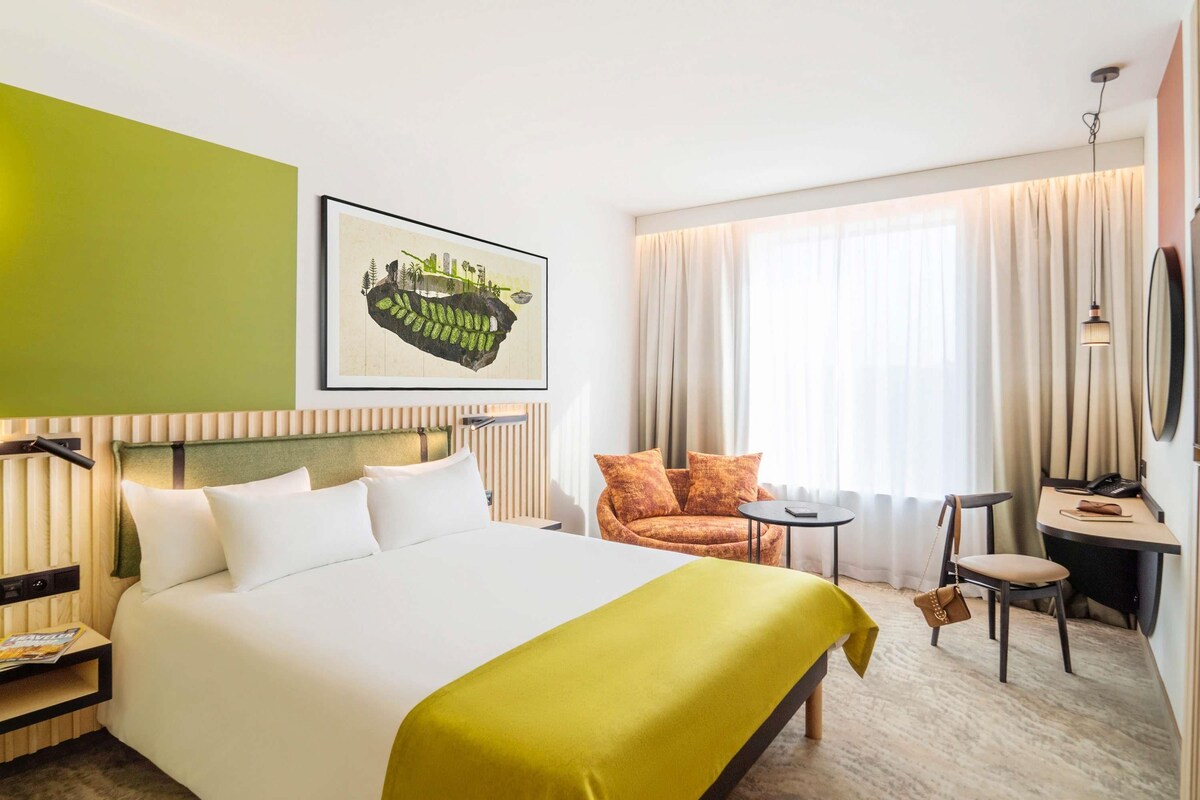 A hotel room with a bed, a chair, a desk, a window with curtains, and a framed picture on the wall.