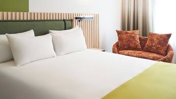 A hotel room with a large bed, a headboard with vertical slats, a sofa with patterned cushions, and a window with curtains.