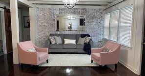 A living room with a brick-patterned wall, a gray sofa, two pink armchairs, a white rug, and a chandelier.