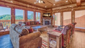 A wooden cabin interior with a fireplace, seating area, and large windows overlooking a forested area. - *Soaring Eagle* VIEWS! Heated POOL-Theater-Game Rm (Sevierville)