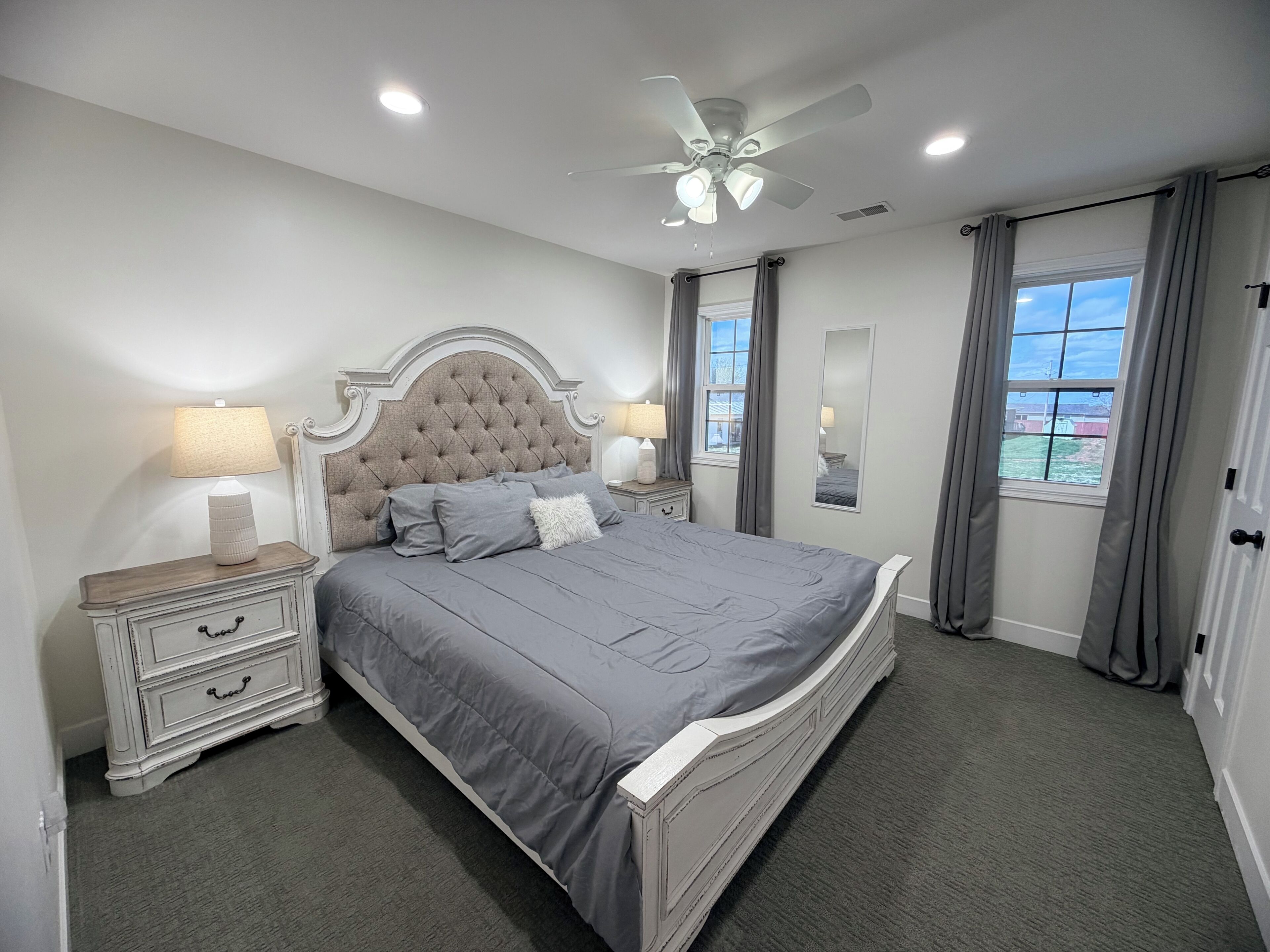Un dormitorio con cama, mesitas de noche, ventilador de techo, ventana con cortinas y vistas al exterior.