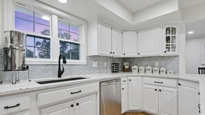 A modern kitchen with white cabinetry, a stainless steel dishwasher, and a window with a view of trees.
