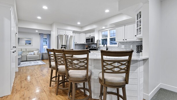 A modern kitchen with white cabinetry, a central island with bar stools, and a dining area with a sofa and a TV.