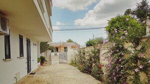 Un vialetto lastricato stretto conduce a un cancello, fiancheggiato da un edificio bianco con persiane e un muro ricoperto di piante rampicanti in fiore.