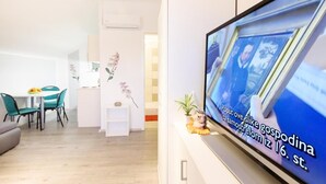 Una sala de estar moderna con un televisor mostrando un noticiero, una zona de comedor con mesa y sillas, y una pared decorativa con una planta.