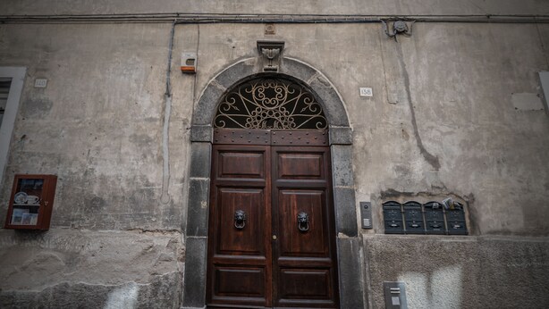 Une grande porte à deux battants en bois sombre, surmontée d’un arc décoratif en métal, est encastrée dans un mur de pierre patiné, orné de plaques numérotées et flanquée d’une petite armoire sur la gauche.