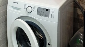 Una lavadora Samsung de carga frontal en un baño con azulejos estampados y un enchufe eléctrico a la vista.
