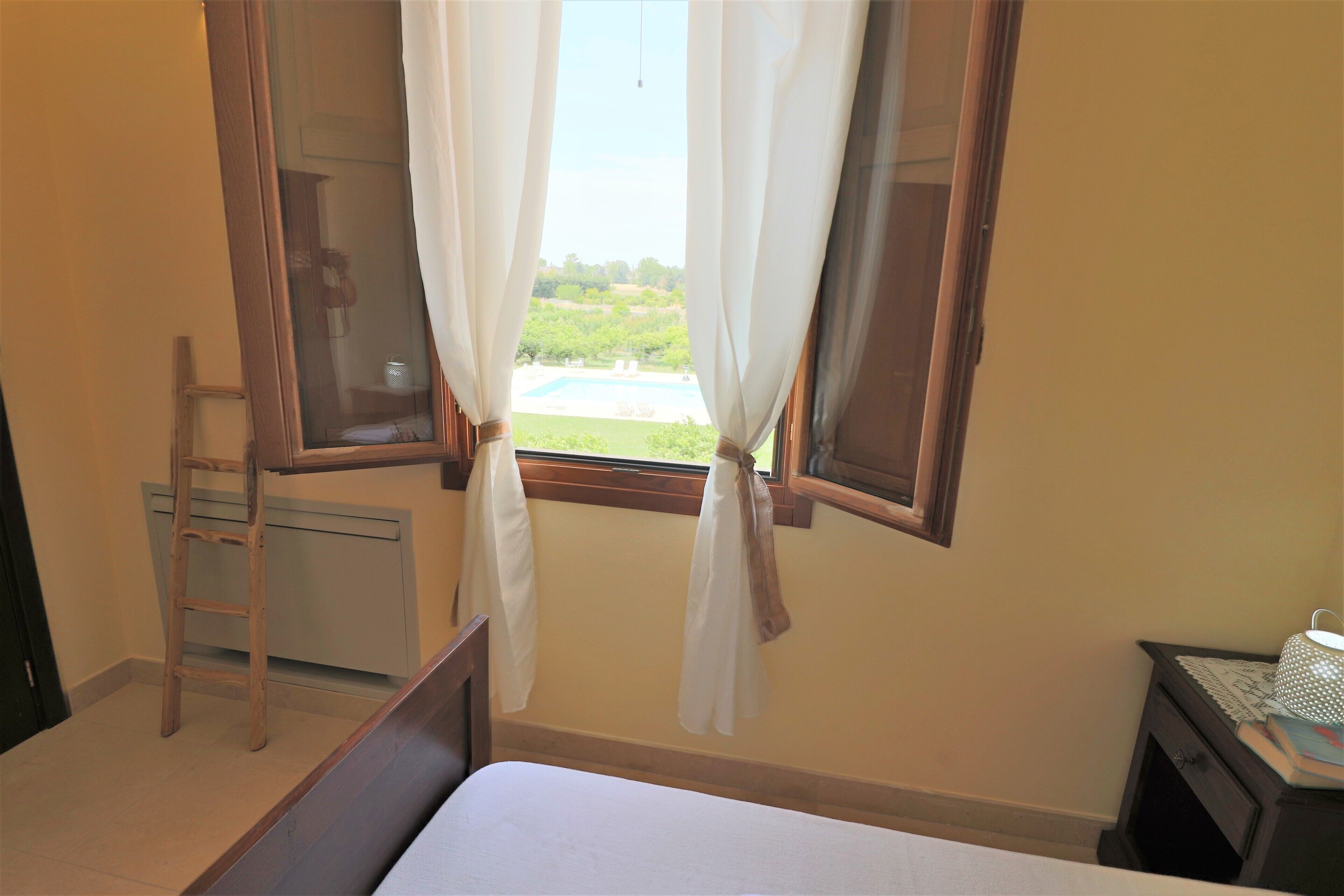 A bedroom with a bed, a window with curtains, a nightstand with a lamp, and a wooden ladder.