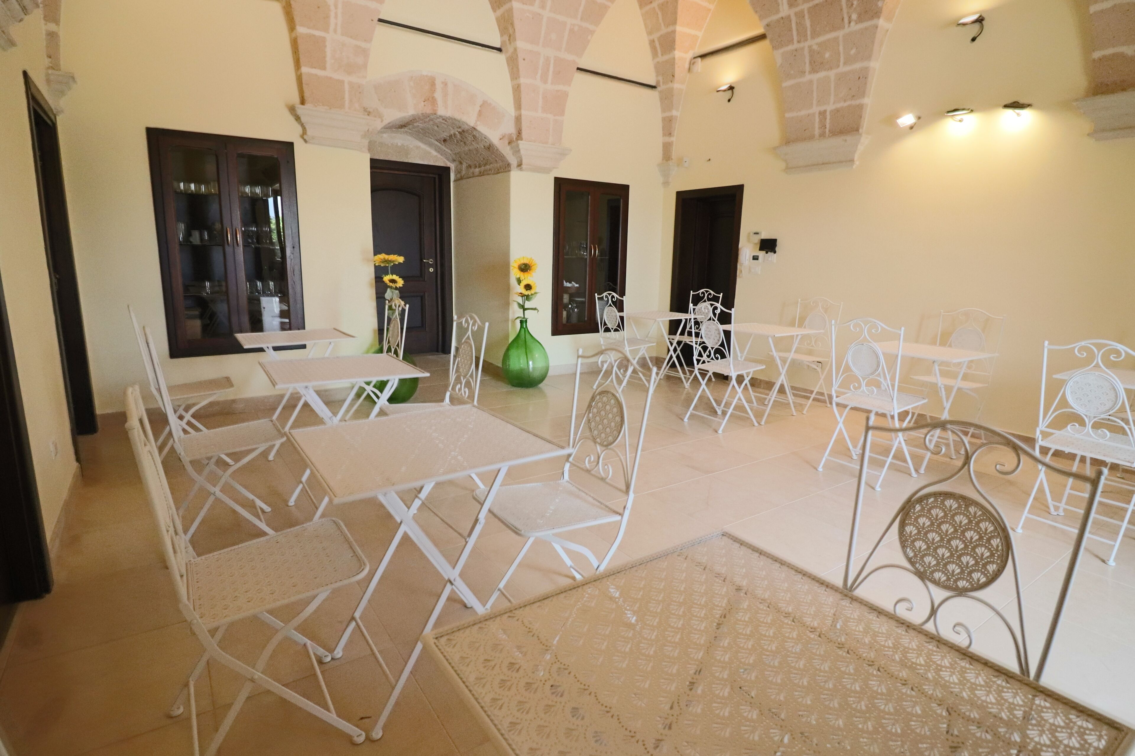A spacious indoor area with white metal tables and chairs arranged in a semi-circle, a green vase with sunflowers, and a stone wall with a door.