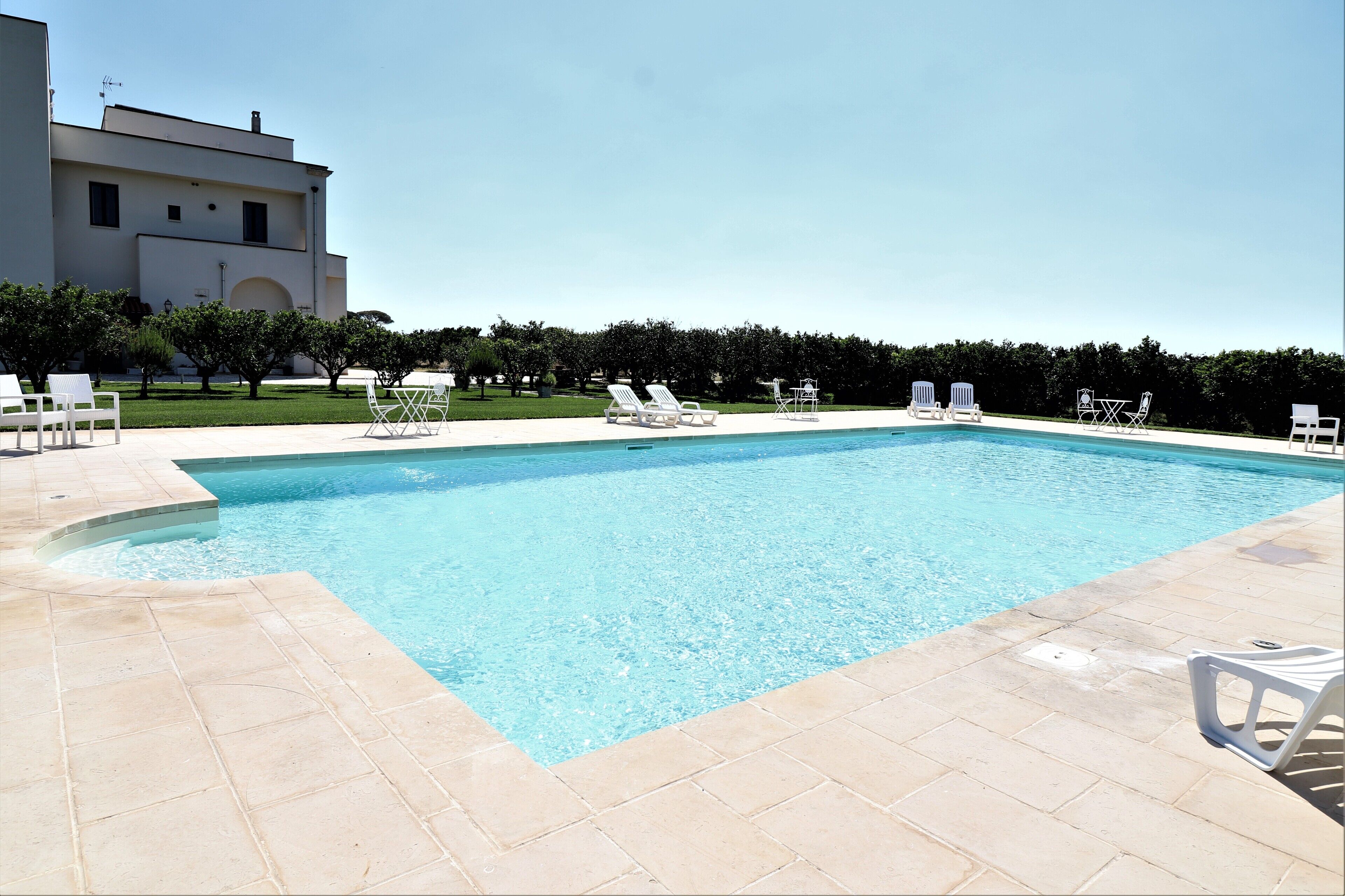 Una piscina rectangular con una solana empaveda, rodeada de sillas de descanso blancas y un césped bien cuidado.