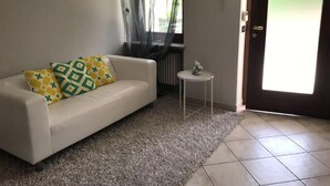 Ein Wohnzimmer mit einem weißen Sofa, gemusterten Kissen, einem kleinen runden Tisch und einer Tür mit Milchglasfenster.