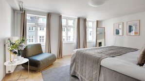 Een moderne slaapkamer met een groot bed, een grijze bank, een kleine tafel met een vaas en meerdere ramen met gordijnen.