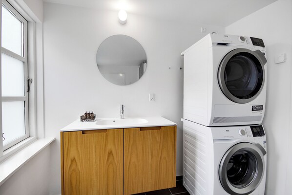 A modern bathroom with a white sink, wooden cabinets, a round mirror, and a stacked washer and dryer. - Regent 3 - Popular 3-bdr Apt With Balcony in Centre (Copenhagen)