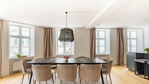 A dining area with a large table, six chairs, a hanging light fixture, and large windows with curtains.