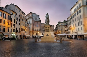 A cobblestone square with historic buildings, a central monument, and outdoor seating areas.
