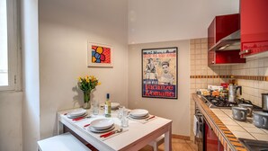 A modern kitchen with a dining area, featuring a white table set with plates, glasses, and cutlery. There are two framed pictures on the wall, a vase with flowers, and a red stove.
