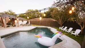 En baghave med swimmingpool, en hvid oppustelig enhjørning, udendørs møbler og lyskæder.
