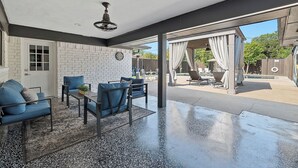 En overdækket terrasse med blåt møbel, en ventilator i loftet og udsigt til en baghave med pergola og udendørs siddepladser.