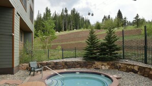 A hot tub surrounded by a stone wall, a wooden deck, and a fenced area with trees and a hill in the background.